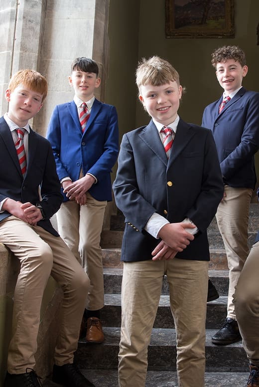 Four boys in blazers, striped ties, and khaki trousers—representing First Year Entry—stand and sit on a staircase, smiling at the camera in a well-lit indoor setting.