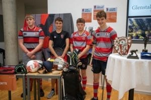 Four teenage boys in red and grey rugby kits stand beside a table displaying sports equipment, trophies, and awards. Indoors, in front of Glenstal signs and a screen, they welcome prospective pupils during Open Day.