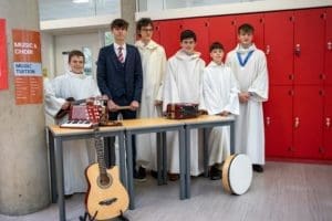 Six boys stand by red lockers and tables with musical instruments, showcasing a vibrant music scene. Perfect for prospective students visiting on Open Day, the Music & Choir sign invites first year students to join the fun.