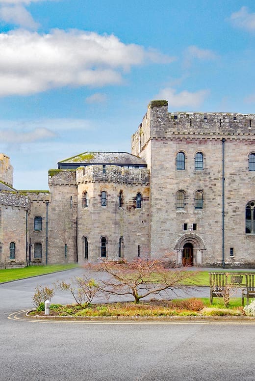 A large stone castle with crenellated towers and arched windows stands under a blue sky with scattered clouds, surrounded by grass and a small landscaped garden—perfect for exploring on your Second Year Entry.